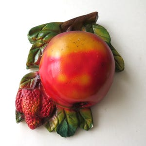 May include: A decorative wall hanging featuring a large red and yellow apple with green leaves and a small cluster of red berries. The apple has a glossy finish and is attached to a brown branch.