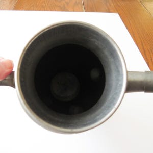 May include: A top-down view of a metal container with a handle and a spout. The interior is dark, and the metal has a dull, silver-gray finish. The container is held by a hand.
