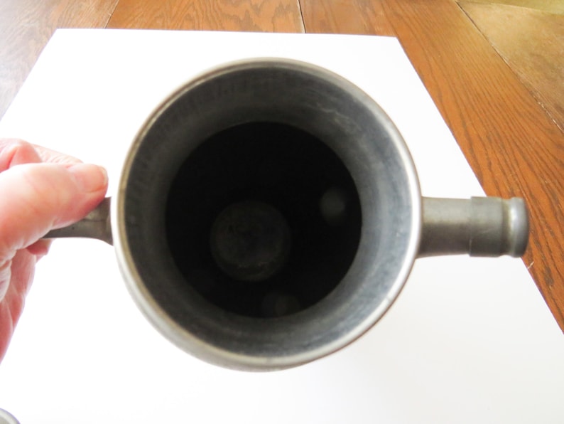 May include: A top-down view of a metal container with a handle and a spout. The interior is dark, and the metal has a dull, silver-gray finish. The container is held by a hand.