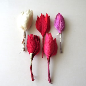 May include: Five artificial flower stems made of layered, pointed feathers in white, red, and purple. Each stem is bound with thin wire, and one red stem features a small white tag with the text MADE IN JAPAN printed in black ink.