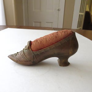 May include: A vintage bronze-colored shoe-shaped pin cushion. The shoe has a pointed toe, decorative detailing, and a small heel. The cushion is a peach-colored fabric, and the shoe is resting on a white surface.