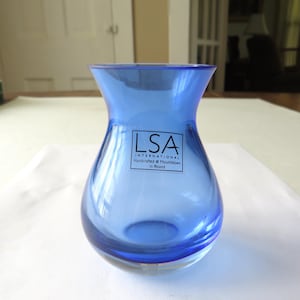 May include: A translucent cobalt blue glass vase with a flared rim and a rounded base. A square white sticker on the front features the black text LSA INTERNATIONAL Handcrafted & Mouthblown in Poland, centered on the body of the glass vessel.