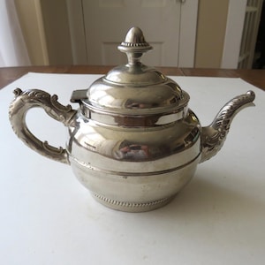 May include: A silver-colored teapot with ornate detailing. The teapot features a curved handle with a dragon-like design, a spout with decorative flourishes, and a domed lid with a finial. The teapot is sitting on a white surface.