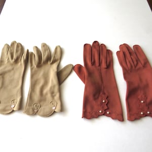 May include: Two pairs of wrist-length gloves, one pair in tan and one pair in rust orange. Each glove features a scalloped cuff edge with decorative detailing. The tan gloves have small buttons, while the rust orange gloves have small beads.