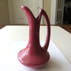 May include: A solid dark pink ceramic pitcher with a smooth, glossy finish. The vessel features a wide, rounded base that tapers into a tall, slender neck with an open, curved spout and a single integrated handle that connects to the upper neck.