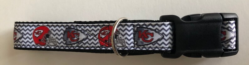 May include: A black and white chevron patterned dog collar with a red and white Kansas City Chiefs logo. The collar has a black plastic buckle.