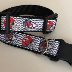 May include: A black and white dog collar with a chevron pattern and red and white Kansas City Chiefs logos. The collar has a black plastic buckle and a silver D-ring.