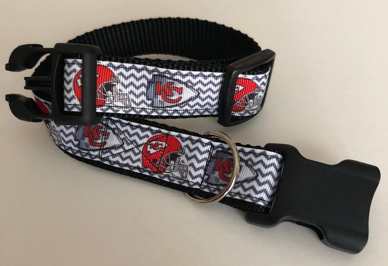 May include: A black and white dog collar with a chevron pattern and red and white Kansas City Chiefs logos. The collar has a black plastic buckle and a silver D-ring.