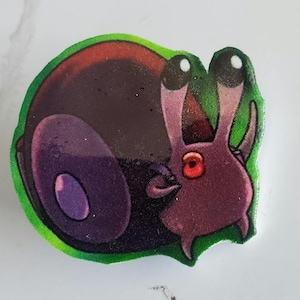 May include: A cartoon-style pin featuring a purple snail-like creature with large, black eyes and a red eye. The pin has a green border and a dark purple shell. The design is whimsical and colorful.