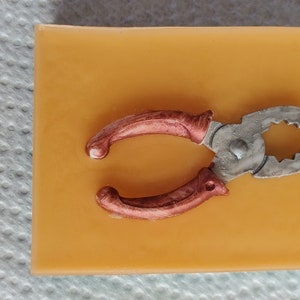 Op de afbeelding: Een tang met rode handvatten op een gele achtergrond. De tang is gemaakt van metaal en heeft een zilveren afwerking.