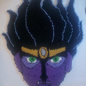 May include: A close-up of a purple and black rug with a face design. The face has green eyes and a gold headband. The rug is made of yarn and has a fuzzy texture.