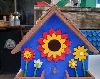 May include: A blue wooden birdhouse with a red, yellow, and white flower design. The birdhouse has two brown planter boxes with green trim.