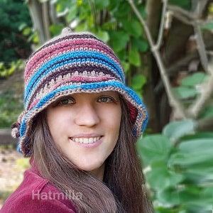 May include: A woman wearing a crocheted hat with brown, pink and blue stripes. The hat has a wide brim and is folded down on one side. The woman has long brown hair and is smiling.