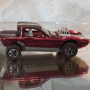 May include: A vintage burgundy toy car with a black roof and silver accents. The car features large wheels with red rims and a detailed engine. The car is reflected in the surface below.