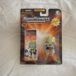 Puede incluir: Un juguete coleccionable Transformers Universe Tiny Titans en su embalaje. El juguete es un robot gris y amarillo que se transforma en un vehículo. El embalaje presenta el logotipo de Transformers Universe y las palabras "Robots in Disguise".