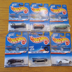 Op de afbeelding: Een verzameling Hot Wheels-speelgoedauto's uit de 1998 First Editions-serie, in hun originele blauw-witte verpakking. De auto's zijn in verschillende kleuren en stijlen, waaronder een zwarte cabriolet, een rode raceauto en een witte sportwagen.