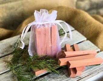 May include: A set of cedar wood blocks in a white organza bag. The blocks are stacked and arranged in a wooden crate with green foliage.