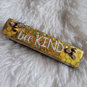 Puede incluir: Un pisapapeles rectangular de resina de color dorado y amarillo con un patrón de panal y dos abejas negras y amarillas. El pisapapeles tiene las palabras "bee kind" escritas en letras blancas.