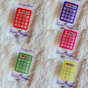 May include: Four colorful calculator-shaped badge reels with the text "I love MATH" and a red heart. The badge reels are on a white background with a white and pink patterned background.