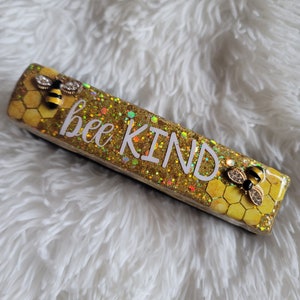 May include: A gold and yellow rectangular sign with glitter and two small bee figurines. The sign says "bee kind" in white letters.
