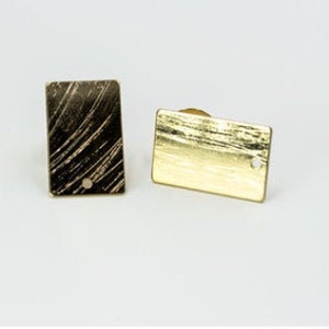 Puede incluir: Dos gemelos rectangulares dorados. Uno tiene un acabado oscuro y texturizado con líneas diagonales, y el otro es de color oro macizo. Cada gemelo tiene un pequeño agujero cerca de la parte inferior.