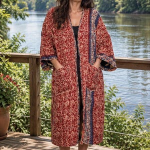 May include: A long, open-front robe featuring a deep red base with a white floral pattern and contrasting blue and white vertical striped panels along the front edges and sleeves. The garment includes two large patch pockets on the front.