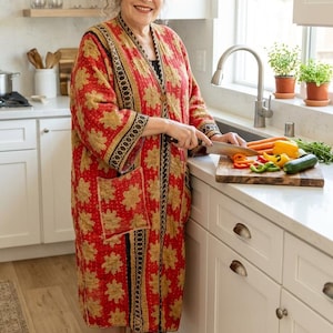 May include: A long-sleeved, knee-length robe in a vibrant red fabric featuring a repeating gold floral pattern. The garment includes a black and white patterned trim along the front opening and cuffs, with a single visible pocket on the front.
