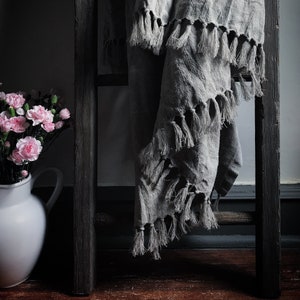 May include: A gray linen throw blanket with fringe detail draped over a dark wooden ladder. A white pitcher filled with pink carnations sits to the left. The scene is set in a dimly lit room, creating a cozy atmosphere.