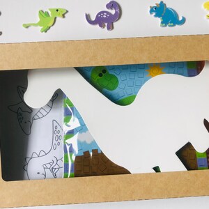 May include: A brown cardboard box with a cutout in the shape of a dinosaur. The cutout reveals a colorful dinosaur-themed design with a white dinosaur silhouette.