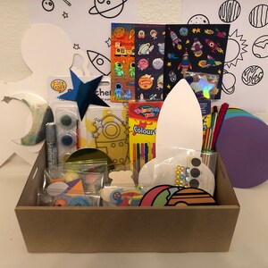 May include: A space-themed craft kit in a cardboard box. It contains a star decoration, paint, stickers, and cut-out shapes of rockets, planets, and stars. The kit also includes colourful pipe cleaners and silver glitter glue.