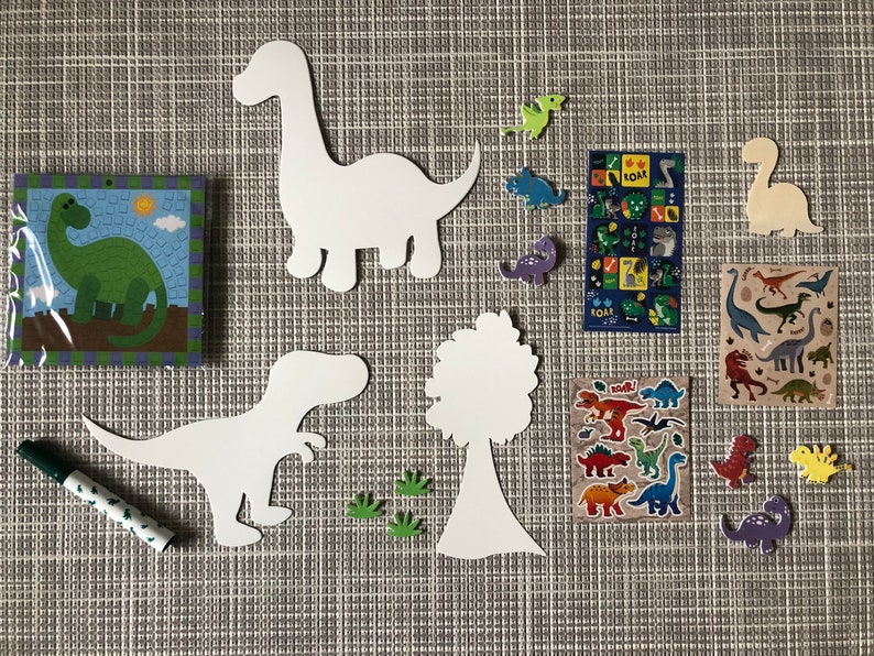 May include: A set of dinosaur-themed craft supplies for kids. The set includes white paper cutouts of dinosaurs, a tree, and footprints, a green marker, and a sheet of dinosaur stickers. The stickers feature colorful dinosaurs with the word "Roar!" printed on them.