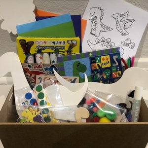 May include: A brown basket filled with dinosaur-themed craft supplies. The basket includes white cardboard dinosaur shapes, colorful craft paper, stickers, crayons, pom-poms, and glue.