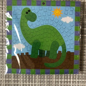 May include: A green dinosaur mosaic craft kit with a blue background, a yellow sun, and white clouds. The kit includes small, colorful squares to create the mosaic.