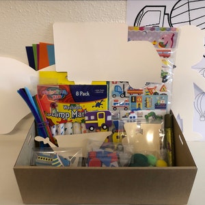 May include: A brown cardboard box filled with craft supplies for kids. The box contains white cardboard shapes of a plane, truck, and hot air balloon, along with colourful pipe cleaners, washable stamp markers, stickers, and foam shapes.