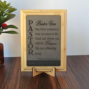 May include: A wooden framed plaque with the text "Pastor Van" and a religious message. The plaque is supported by a wooden stand and is displayed on a dark wooden surface. The text is in a dark color.