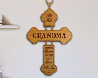 May include: A light brown wooden wall hanging composed of three connected pieces. The top piece features a sun icon, the middle piece displays the word "GRANDMA," and the bottom piece contains the text "You're the Sunshine that helps me to Grow I Love You Love, Charley."