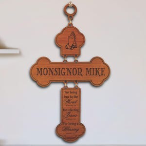 May include: A brown wooden wall hanging cross featuring a top section with praying hands, a center horizontal bar engraved with the text MONSIGNOR MIKE, and a vertical bottom section with engraved religious verses and scripture references.