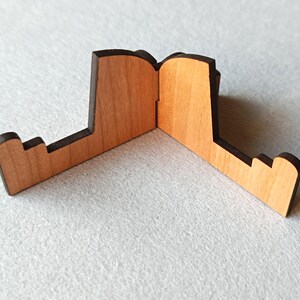 May include: A wooden phone stand with a natural wood finish and dark brown edges. The stand is designed with a unique shape, featuring a central hinge and two angled support arms. The stand is designed to hold a phone or small tablet.