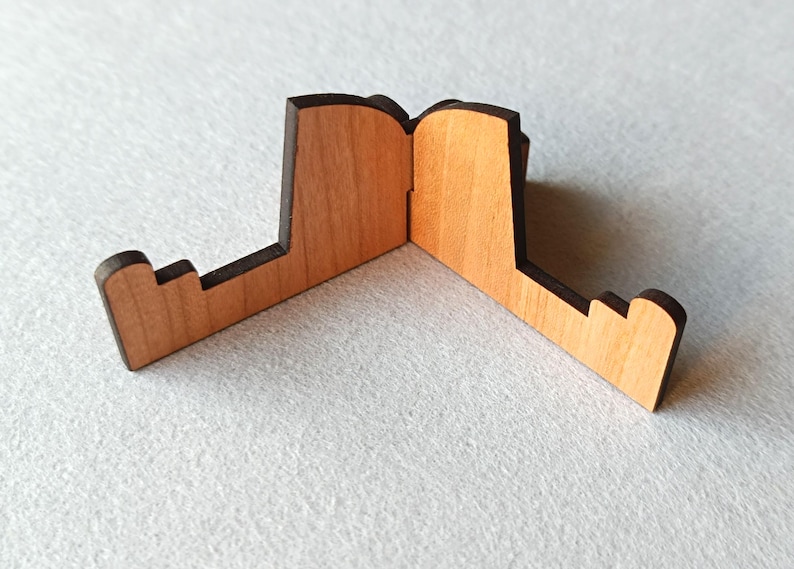 May include: A wooden phone stand with a natural wood finish and dark brown edges. The stand is designed with a unique shape, featuring a central hinge and two angled support arms. The stand is designed to hold a phone or small tablet.