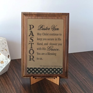 May include: A wooden framed plaque with a message for Pastor Van. The text reads "May Christ continue to keep you secure in His Hand, and shower you with His Grace. You are a Blessing to us." The plaque is supported by a wooden stand.