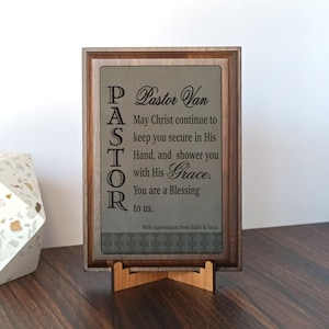 May include: A wooden framed plaque with the word "PASTOR" vertically on the left side. The plaque has a light gray background with the text "Pastor Van" and a religious message. It is displayed on a wooden stand.
