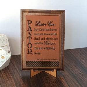 May include: A wooden framed plaque with a brown leather-like insert. The text reads "Pastor Van" and a religious message. The plaque is supported by a wooden stand. The text "PASTOR" is written vertically on the left side.