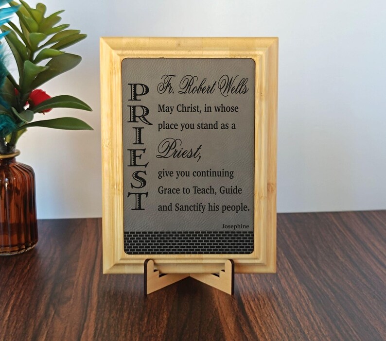 May include: A framed wooden plaque with the word "PRIEST" vertically aligned on the left side. The plaque features a gray background with text that reads "Fr. Robert Wells" and a prayer. The frame is light brown, and the plaque is displayed on a wooden stand.