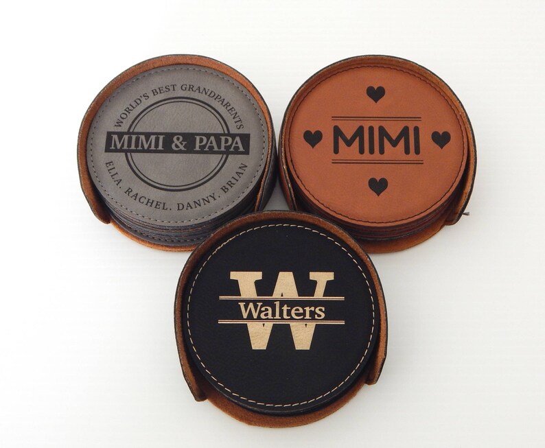 May include: Set of three leather coasters with personalized engravings. The first coaster is gray with the text "World's Best Grandparents Mimi & Papa Ella Rachel Danny Brian". The second coaster is brown with the text "Mimi" and two hearts. The third coaster is black with the text "Walters" and a letter W.