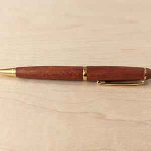 May include: A ballpoint pen featuring a polished reddish-brown wood barrel with gold-tone metal accents, including a tapered tip, a center band, and a pocket clip. The pen has a smooth, rounded end and a visible metal writing point at the front.