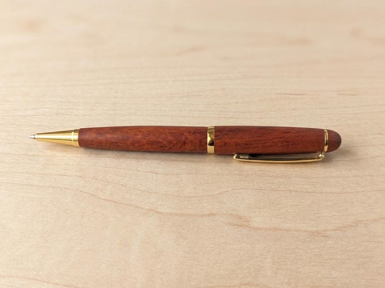 May include: A ballpoint pen featuring a polished reddish-brown wood barrel with gold-tone metal accents, including a tapered tip, a center band, and a pocket clip. The pen has a smooth, rounded end and a visible metal writing point at the front.