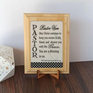 May include: A wooden framed sign with the word "PASTOR" vertically aligned on the left side. The sign includes the text "Pastor Van, May Christ continue to keep you secure in His Hand, and shower you with His Grace. You are a Blessing to us."