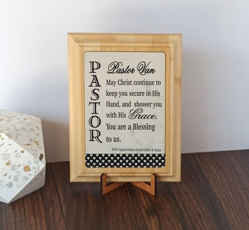 May include: A wooden framed sign with the word "PASTOR" vertically aligned on the left side. The sign includes the text "Pastor Van, May Christ continue to keep you secure in His Hand, and shower you with His Grace. You are a Blessing to us."