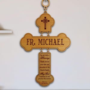May include: A light brown wooden wall hanging composed of three connected pieces. The top piece features a cross, the middle piece reads FR. MICHAEL, and the bottom piece contains a message of gratitude and blessing with the signature With love, The Edwards.