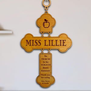 May include: A light brown wooden wall hanging shaped like a cross, featuring a chef hat icon with an apple, the name MISS LILLIE, and the engraved text: To TEACH is to TOUCH lives forever. Thank you for caring. Nina Torres & Family.